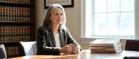 Senior female attorney with gray hair in professional office setting, seated at desk with law books and documents, wearing blazer, contemplative expression, suitable for legal services, law firm marketing, and professional business content.