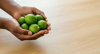 Person with dark skin tone holding fresh green limes in cupped hands over wooden surface. Suitable for food, health, cooking, and organic produce concepts.