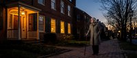 Elderly man with walking cane strolls past historic brick townhouses at twilight on cobblestone street with warm glowing windows creating cozy neighborhood atmosphere.
