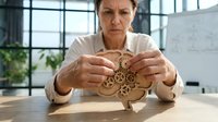 Focused middle-aged Caucasian businesswoman assembling wooden gear puzzle at office desk, problem-solving and strategic thinking concept for corporate training.