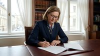 Mature woman with light brown hair and fair complexion, wearing glasses and dark blazer, writing at wooden desk in elegant office with bookshelves and windows.