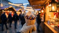 Hands holding roasted chestnuts in paper cone at festive Christmas market with warm lights and wooden stalls creating cozy winter atmosphere.