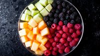 Fresh colorful fruit bowl with cubed melon, pineapple, blackberries and raspberries arranged in sections on dark background, top view food photography.