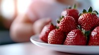 Fresh ripe strawberries on white ceramic plate with soft blurred background. Close up view shows detailed texture and natural shine of bright red berries.
