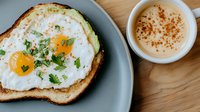 Sunny side up eggs on toasted bread garnished with fresh herbs and spices served with creamy coffee latte dusted with cinnamon on wooden table. Morning breakfast setting.