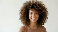 Young African American woman with natural curly afro hair smiling warmly at camera wearing brown top against neutral background for lifestyle content.