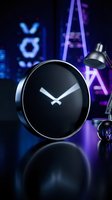 Modern black round clock on reflective surface with neon purple and blue lighting in contemporary office workspace for time management concepts.