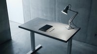 Modern minimalist workspace with adjustable desk, smartphone, and black task lamp in contemporary office interior with concrete walls and natural lighting.