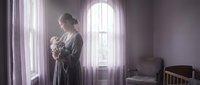 Young Caucasian mother in gray robe holding newborn baby by large window in soft purple nursery room with natural light streaming through curtains.