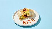 Breakfast burrito cut in half on a white plate with hot sauce spelling BITE, served on a light blue background.