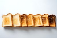 Seven slices of white bread arranged in row showing gradual toasting progression from lightly toasted to dark burnt, displayed on light background.