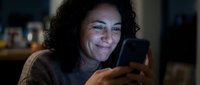 Middle-aged woman with dark curly hair and olive complexion, wearing a brown sweater, smiling while holding and looking at a smartphone in an indoor setting with warm lighting.