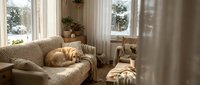 Cozy living room interior with orange tabby cat sleeping on beige sofa near large windows with natural sunlight streaming in during winter day.