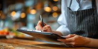 Server in striped apron taking order on clipboard in restaurant with warm ambient lighting and blurred colorful background of food displays.