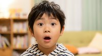 Surprised Asian boy with wide eyes and open mouth expressing shock or amazement in bright home interior with books and couch in background.
