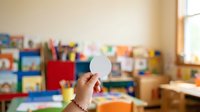 Child's hand holding blank white circle paper against colorful classroom background with educational toys and supplies, suitable for learning concepts, creativity projects, and school communications.