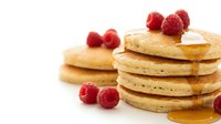 Golden fluffy pancakes stacked high with fresh raspberries and maple syrup drizzling down, breakfast comfort food on white background isolated.