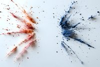 Red and blue watercolor paint splashes facing each other on white paper, creating dynamic abstract explosion effect with scattered droplets and splatters.