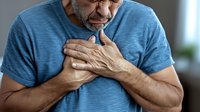 Middle-aged Caucasian man in blue t-shirt experiencing chest pain, clutching heart area with both hands, showing signs of possible cardiac distress or heart attack symptoms.