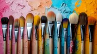 Makeup brushes arranged in row against colorful textured background with pink, turquoise and orange paint strokes, showcasing professional cosmetic tools and artistry.