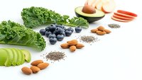 Healthy superfood ingredients arranged in row including kale, blueberries, avocado, almonds, chia seeds and carrots on white background for clean eating.