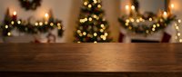 Empty wooden table surface with blurred Christmas tree and warm golden lights in cozy holiday living room background for product display.