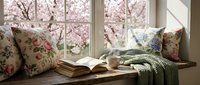 Cozy reading nook with floral cushions, open book and knitted throw blanket by window overlooking blooming cherry blossom trees in spring sunlight.