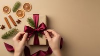 Female hands tying burgundy ribbon on kraft paper gift box with dried orange slices, cinnamon sticks, star anise and pine branches on beige background.