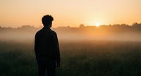 Silhouette of young man standing in misty field at golden sunrise. Contemplative moment in nature with warm morning light and peaceful countryside atmosphere.