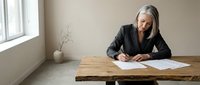 Mature Asian businesswoman with gray hair writing documents at wooden desk in minimalist office space with natural lighting for professional content.