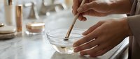 Woman mixing skincare ingredients in glass bowl with wooden spoon for homemade natural beauty treatment preparation in modern bathroom setting.