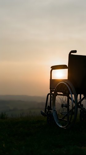 Wheelchair silhouetted against golden sunset sky on grassy hilltop overlooking misty valley landscape. Mobility aid symbolizing freedom, hope, and independence during golden hour.
