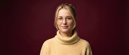 Young blonde Caucasian woman in yellow turtleneck sweater and glasses smiling confidently against burgundy background for professional headshot.