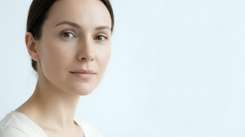Professional portrait of confident young Caucasian woman with natural makeup and brown hair pulled back, wearing white top against clean background.