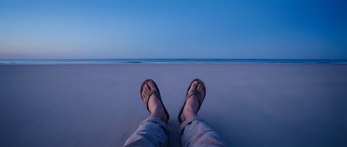 Young adult male wearing casual sandals and blue jeans sitting at edge of calm ocean water at dusk, feet dangling over serene seascape horizon.