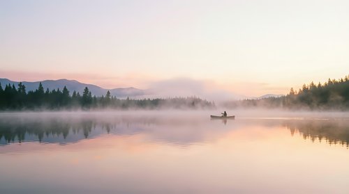 Serene lake at sunrise with mist rising from calm water, lone fisherman in boat, forested shoreline, peaceful morning atmosphere, soft pink and purple sky reflecting on glassy surface.