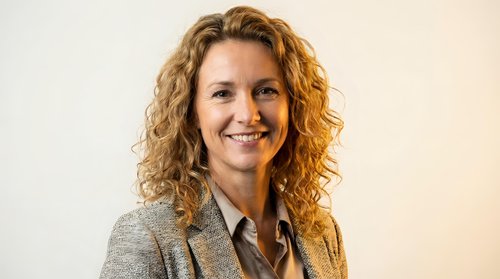 Professional middle-aged Caucasian businesswoman with curly blonde hair smiling confidently in gray blazer against neutral background for corporate use.