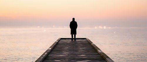 Silhouetted male figure standing alone on wooden pier at sunrise, gazing toward calm ocean horizon during golden hour, conveying solitude, contemplation, and peaceful morning reflection.