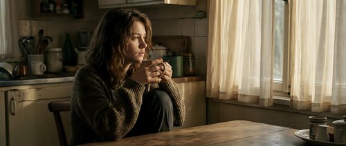 Young Caucasian woman in cozy knit sweater sitting at wooden kitchen table, holding coffee mug, gazing thoughtfully out window in warm morning light.