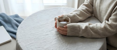 Woman in cozy beige knit sweater holding steaming mug at table, soft natural light, warm hygge atmosphere for lifestyle and wellness content.