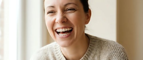 Happy middle-aged Caucasian woman laughing joyfully in cozy beige sweater at home, expressing genuine happiness and positive emotions indoors.