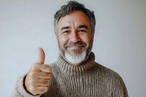 Mature bearded man in cozy knit turtleneck sweater showing thumbs up gesture with positive friendly smile against light wall background.