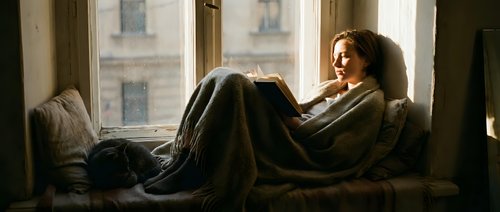 Young woman relaxing on couch wrapped in cozy blanket by window with natural light creating peaceful morning atmosphere for wellness and comfort concepts.