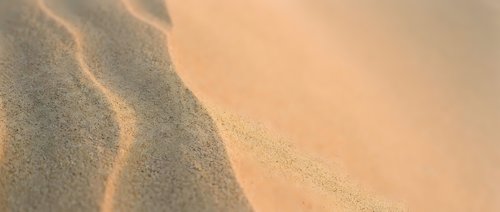 Young adult male with light skin tone wearing casual beige clothing, reclining on sandy beach with warm golden sunlight creating soft shadows across textured sand surface.