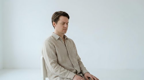 Young Caucasian man in beige linen shirt sitting thoughtfully against white background, minimalist portrait for lifestyle and fashion content.