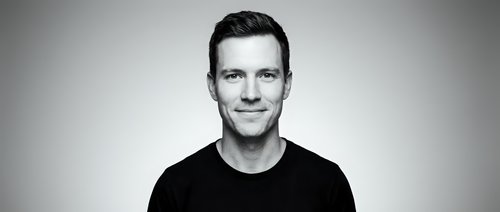 Portrait of a young adult male with fair complexion and dark hair, wearing a black casual t-shirt against neutral background for professional headshots and corporate use.