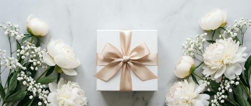 White gift box with beige ribbon surrounded by white roses and baby's breath flowers on marble background for special occasions and celebrations.