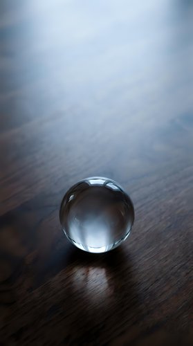 Clear crystal glass sphere resting on dark wooden surface with soft blue light, casting a delicate shadow in a moody, minimalist composition.