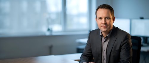 Professional middle-aged Caucasian businessman in dark suit sitting at modern office desk with confident smile for corporate presentations and marketing.