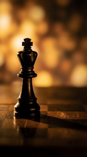 Black chess king piece on wooden board with warm golden bokeh background creating dramatic lighting for strategy and leadership concepts.